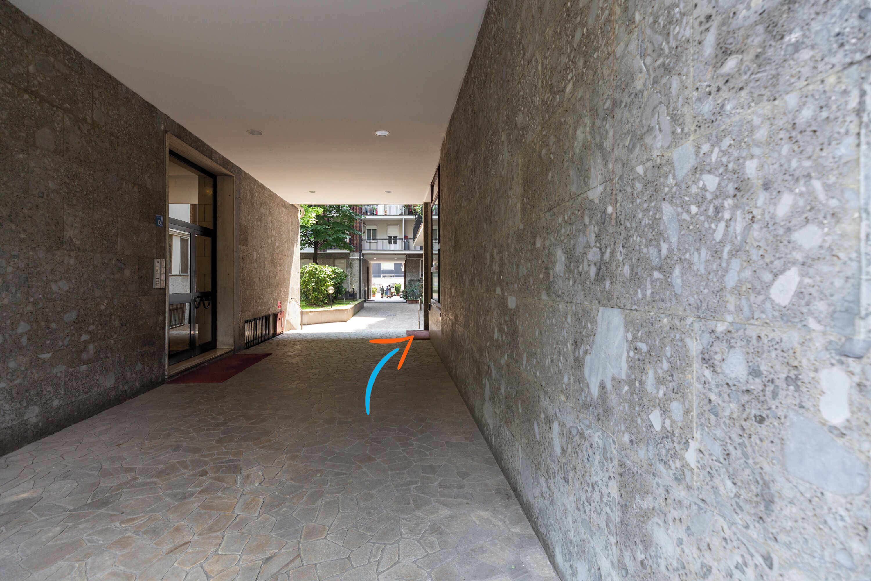 A corridor with stone walls and a tiled floor, leading to an open area with a view of buildings in the distance.
