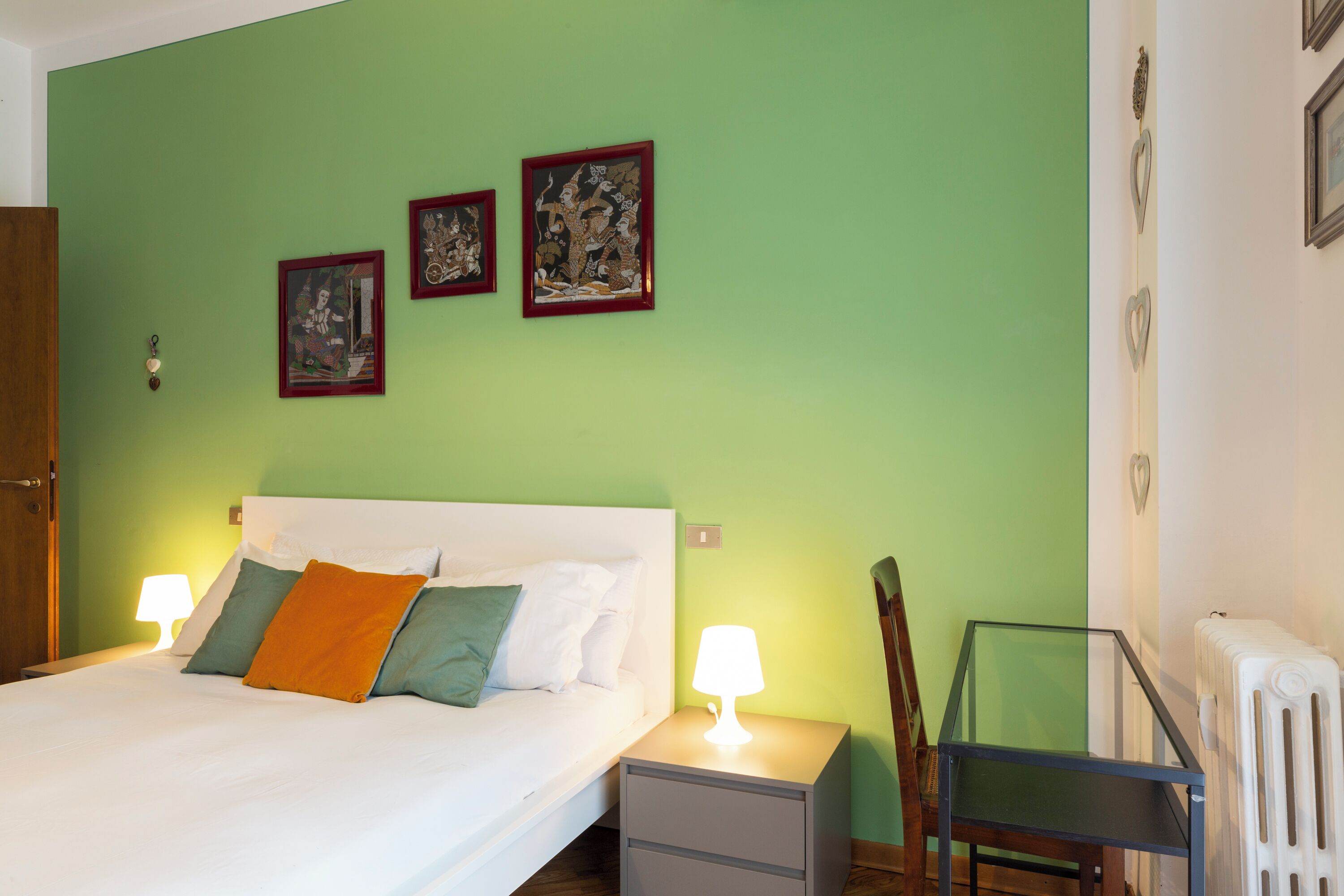 A bedroom with a bed, two bedside lamps, a nightstand, a chair, and three framed artworks on the wall.