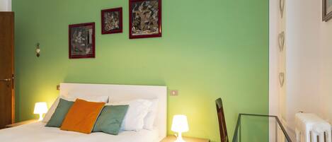 A bedroom with a bed, two bedside lamps, a nightstand, a chair, and three framed artworks on the wall.