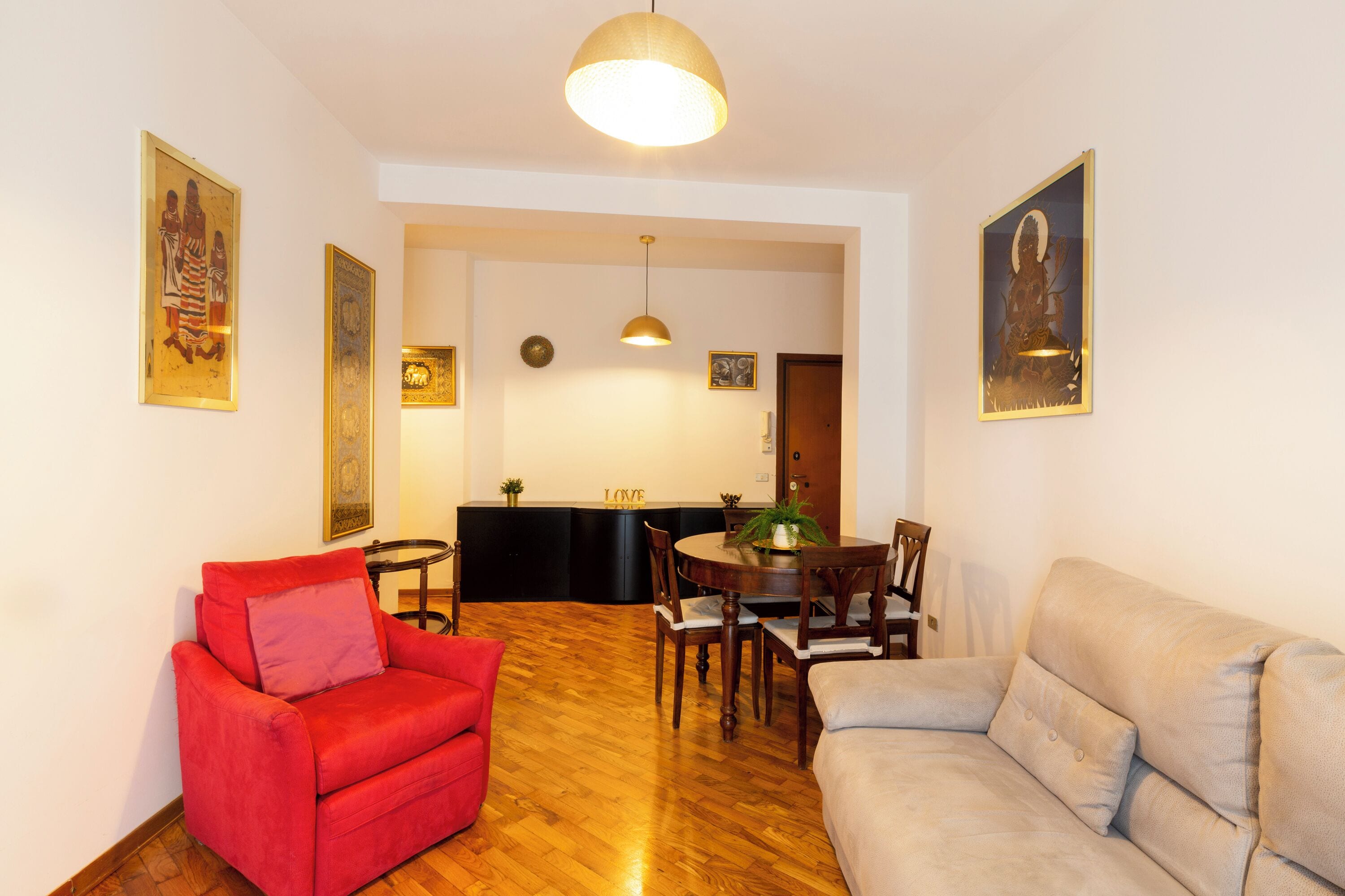 A living room with a red armchair, a beige sofa, a wooden dining table with chairs, and a kitchen in the background.