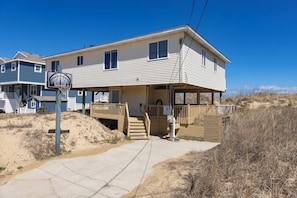 A two-story house with a covered porch, a basketball hoop, and a sandy beach in the background. - An Octopus Hug, Oceanfront, Dog Friendly, 4 Bd. (Virginia Beach)