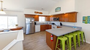 Una cocina moderna con armarios de madera, un refrigerador de acero inoxidable, un microondas y un lavamanos. Hay taburetes de bar verdes y un extintor de incendios montado en la pared.