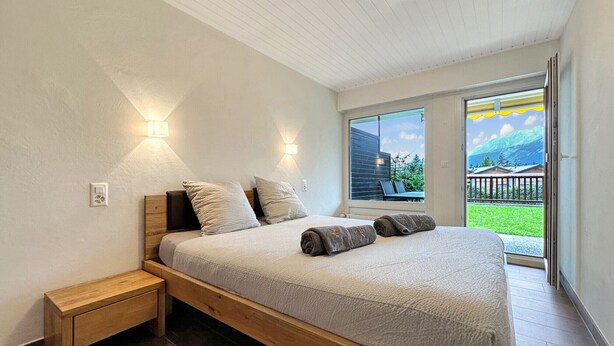 Un dormitorio con cama, mesita de noche y ventana con vista a una casa y montañas.
