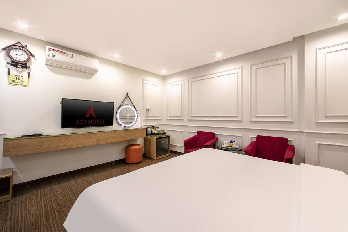Una habitación de hotel con una cama grande, televisión, reloj, aire acondicionado y una pequeña mesa con una silla roja.