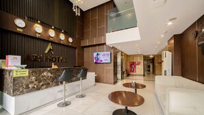 Ein Hotel-Rezeptionsbereich mit einer Marmor-Theke, zwei Barhocker, einem Fernsehbildschirm und einer Sitzecke mit einer Couch und runden Tischen.