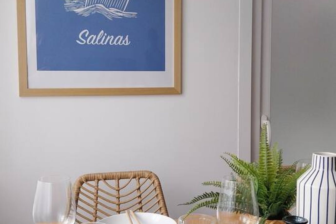 A framed artwork with the word 'Salinas' on a blue background, a wicker chair, a glass table with a white plate, a glass, a striped vase, and a fern plant.
