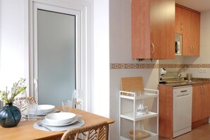 A dining area with a wooden table set for two, a white cabinet, and a kitchen area with a dishwasher. - 11A02 Casa RTPG  6p (Castrillon)