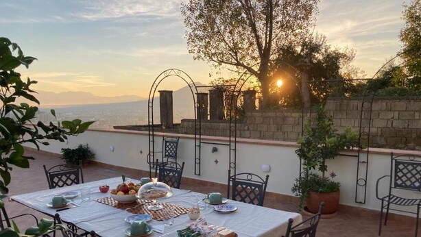 Un área de comedor al aire libre con una mesa dispuesta para dos personas, rodeada de sillas, y vista al mar y a las montañas de fondo.