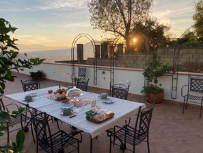 An outdoor dining area with a table set for two, surrounded by chairs, with a view of the sea and mountains in the background. - Le Petit Palais Naples (Naples)