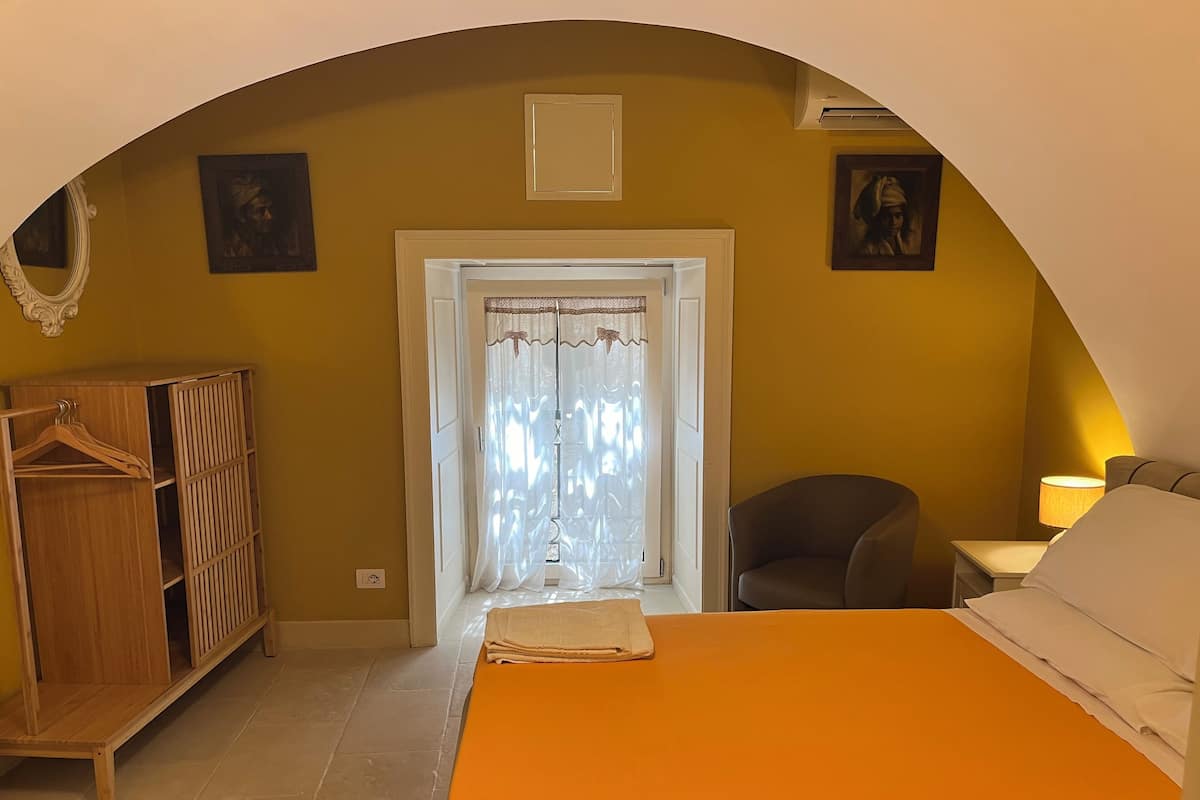 A bedroom with an arched doorway, a wooden wardrobe, a bed with an orange bedspread, a chair, and a nightstand with a lamp.