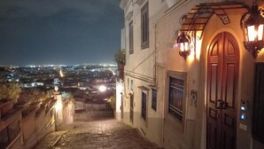 Vista nocturna de una escalera de piedra que conduce a un edificio con puerta de madera y ventanas arqueadas, con vistas a un paisaje urbano iluminado.