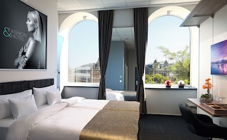 Quarto de hotel com cama, escrivaninha e uma grande janela com cortinas.