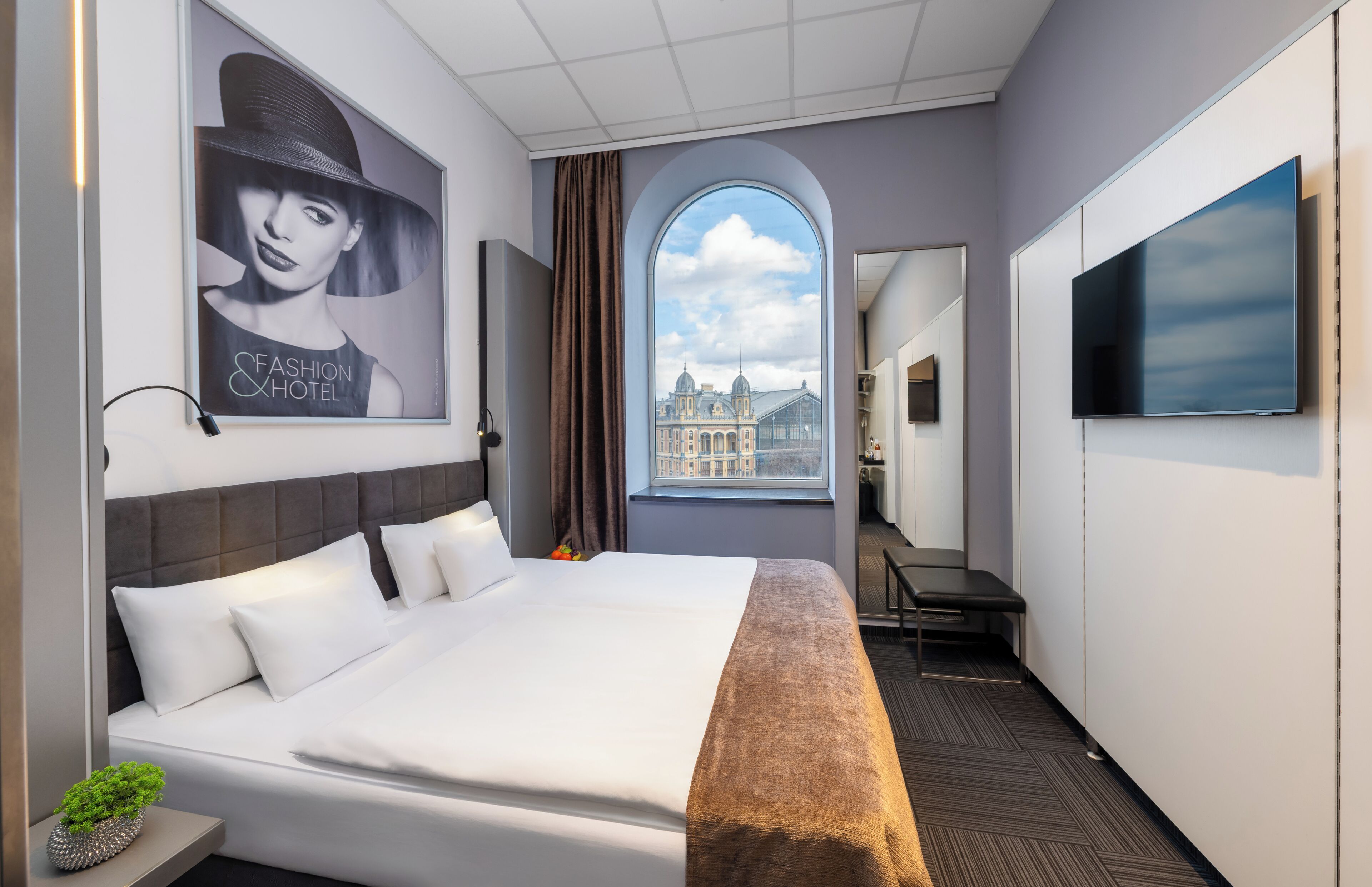 A hotel room with a large bed, a TV, a window with a view of a building, and a poster on the wall.