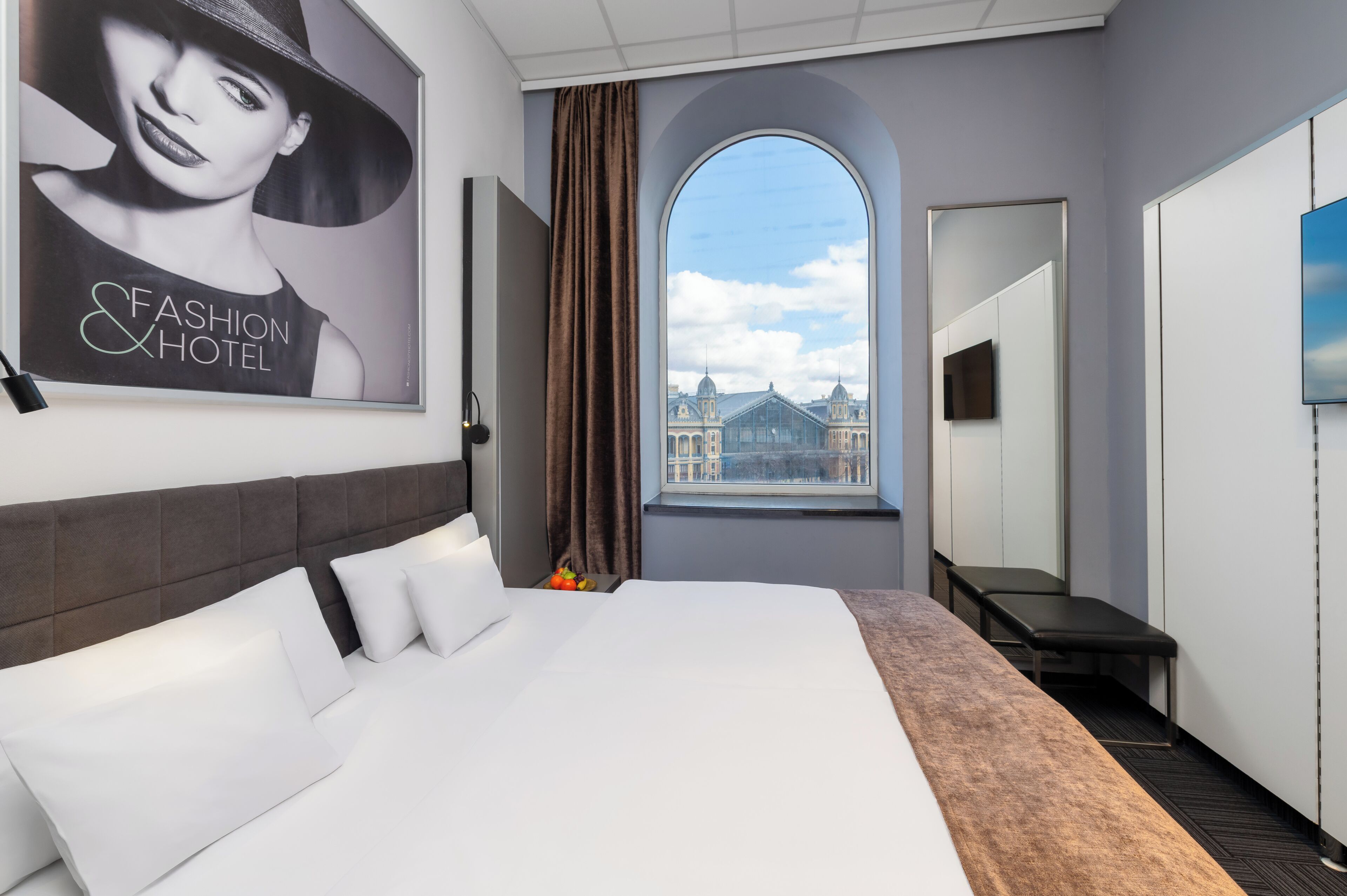 Habitación de hotel con una cama grande, vista a un edificio por la ventana, un televisor de pantalla plana y un espejo.