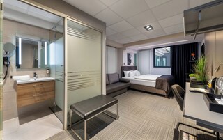 Moderni hotellihuone, jossa on lasiseinä, suuri sänky, penkki, työpöytä sekä kylpyhuone, jossa on peili ja pesuallas.