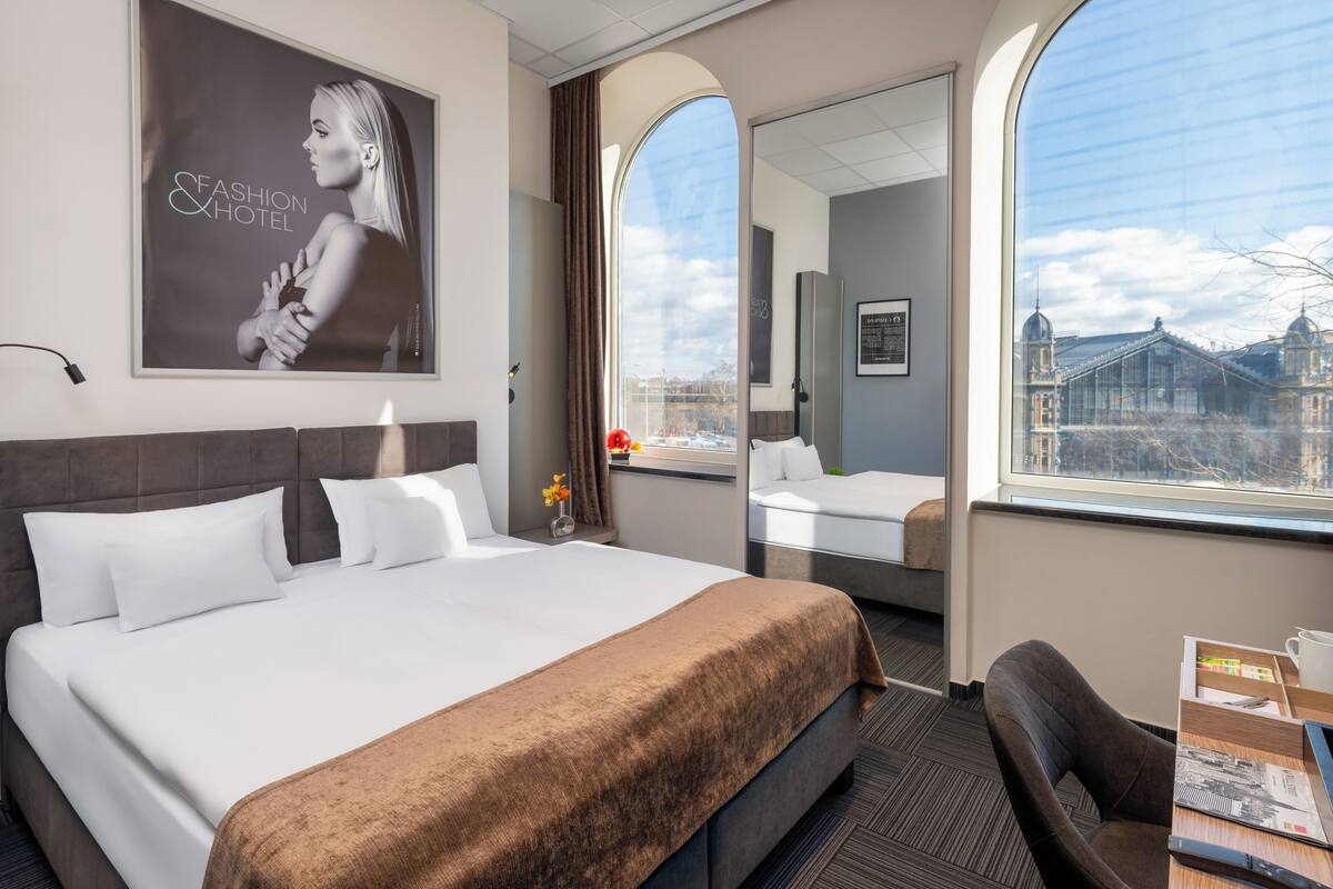 A hotel room with a large bed, a desk, a chair, a window with a city view, and a poster on the wall.