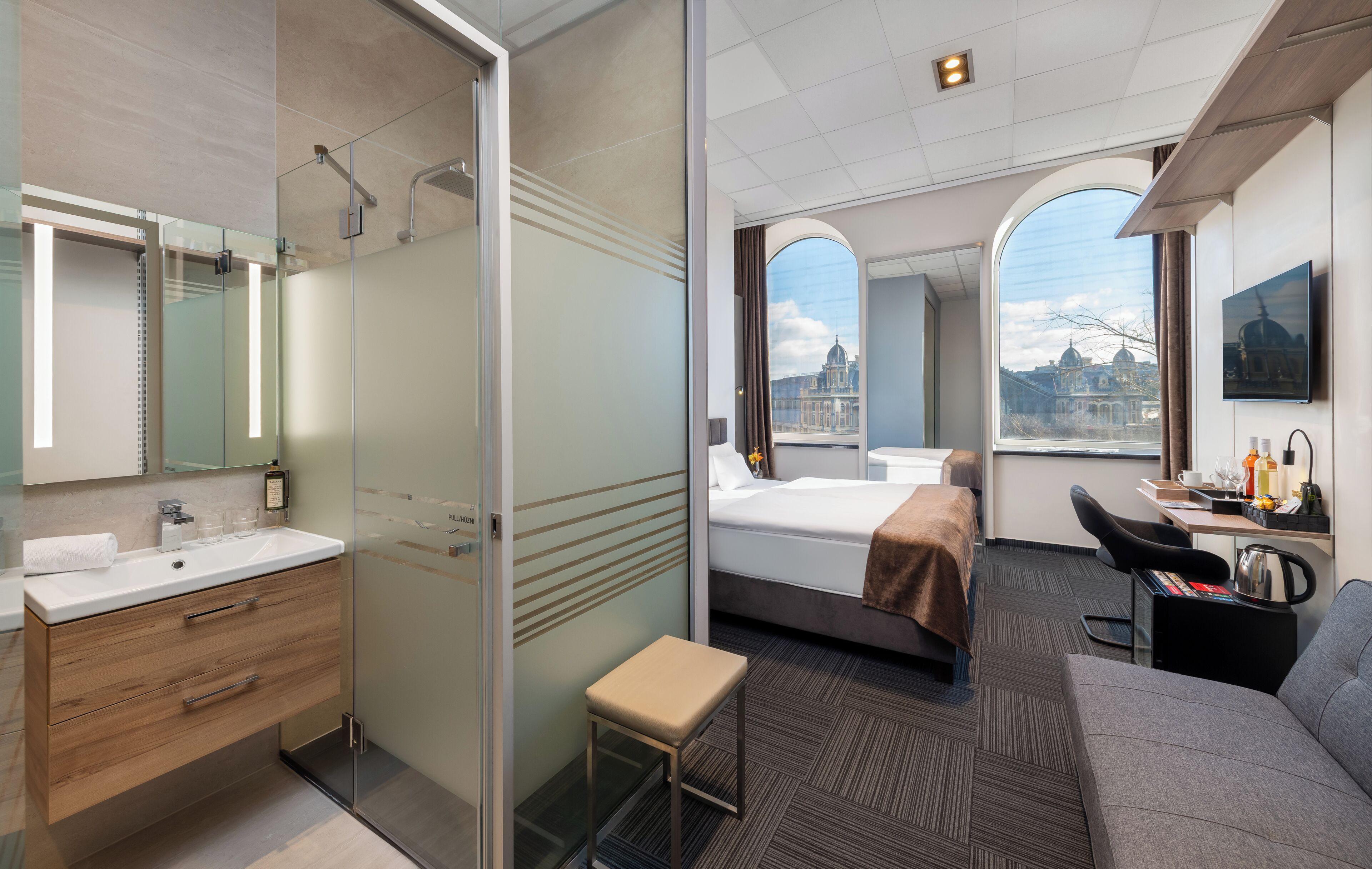Een moderne hotelkamer met een glazen douche, een bed, een bureau en uitzicht op een stadsbeeld.