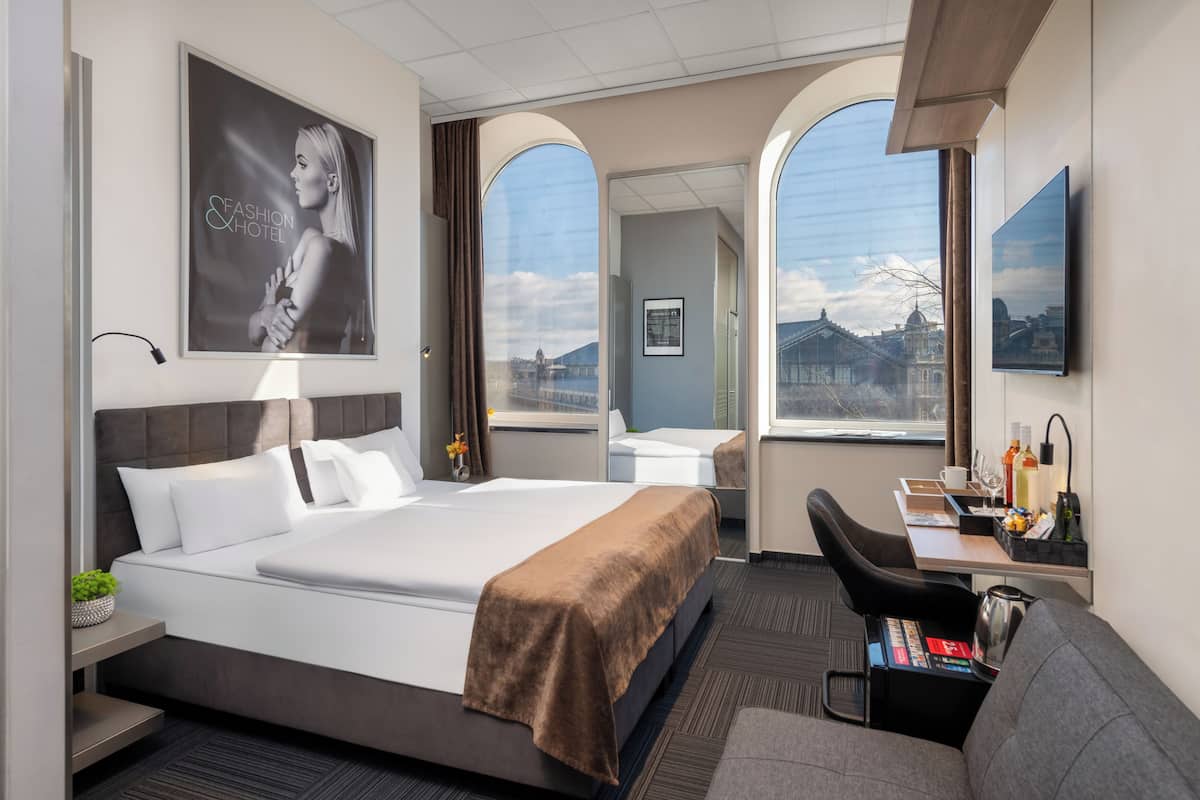 A hotel room with a large bed, a desk with a chair, a TV, and a view of a cityscape through arched windows.