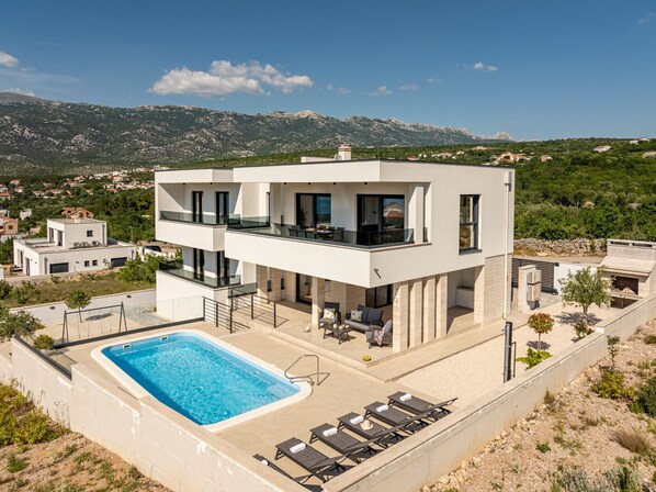 A modern two-story house with a swimming pool, outdoor seating, and a view of the surrounding landscape.