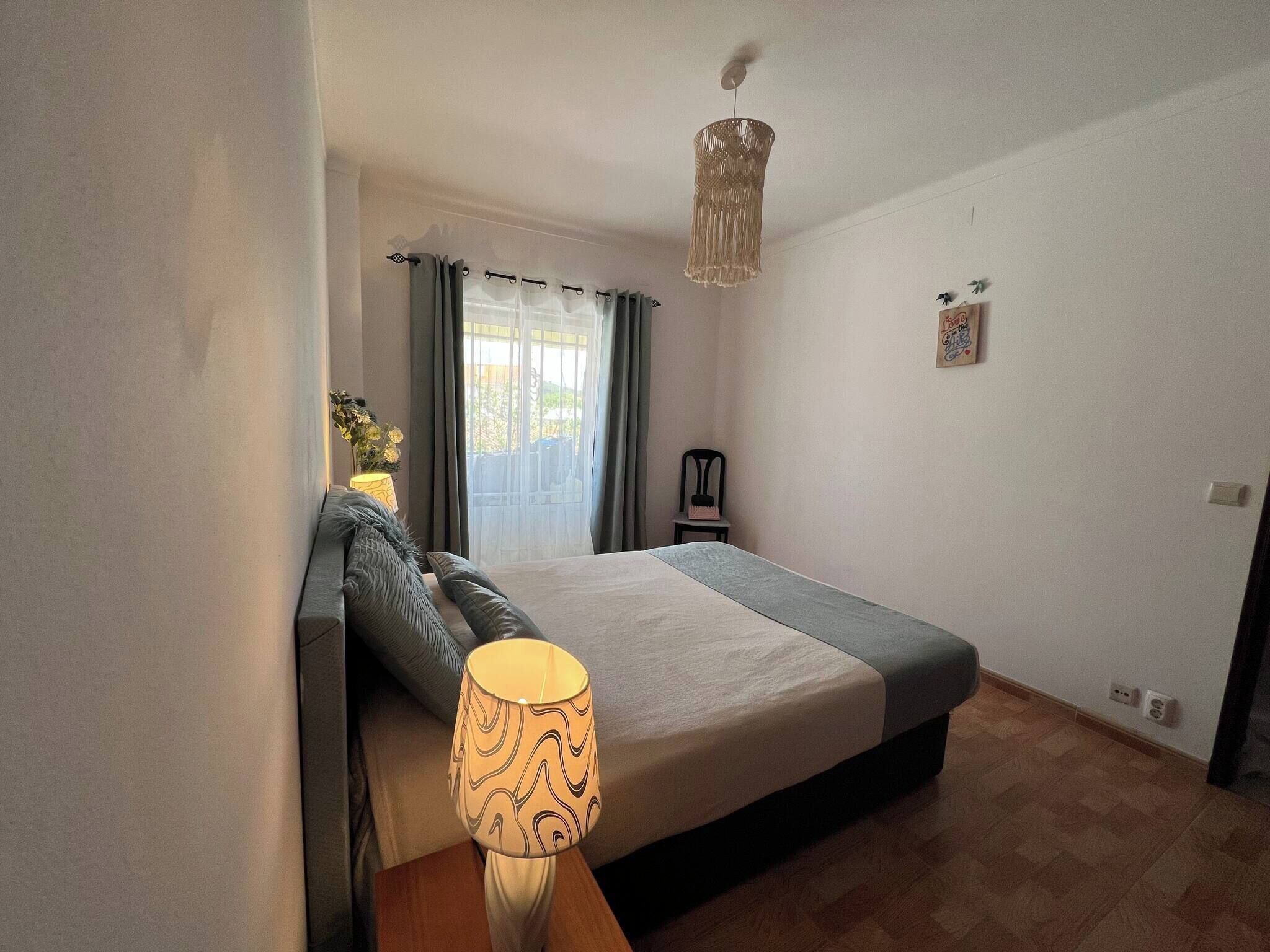 Un dormitorio con cama, lámpara de noche, sillón, ventana con cortinas y una lámpara colgante.