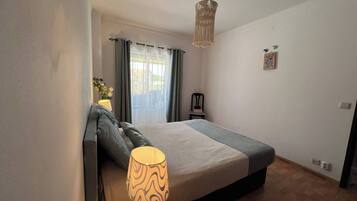 Un dormitorio con cama, lámpara de noche, sillón, ventana con cortinas y una lámpara colgante.
