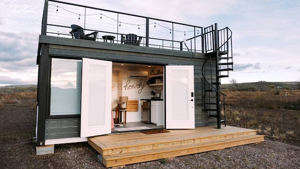 Une maison moderne et compacte, dotée d’un balcon, d’un escalier et d’une cuisine visible à travers une porte ouverte.