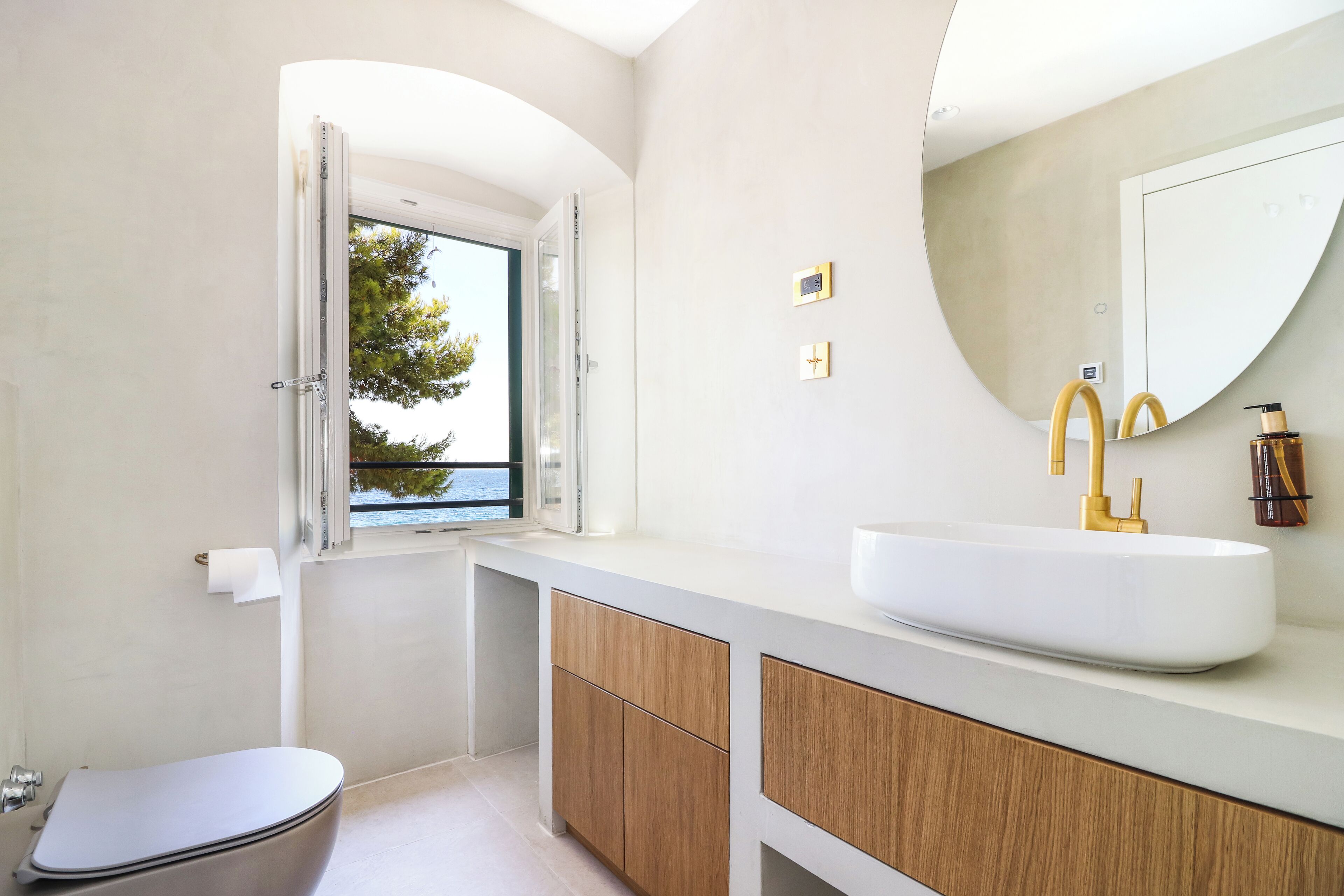 A modern bathroom with a toilet, a sink with a gold faucet, wooden cabinets, and a large mirror.