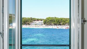 A room with a large window offering a view of a beach, ocean, and buildings in the distance. - Vila Fenč (Primosten)