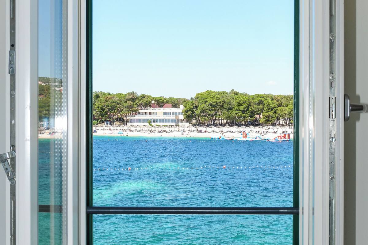 Habitación con un amplio ventanal que ofrece vistas a una playa, el océano y edificios al fondo.