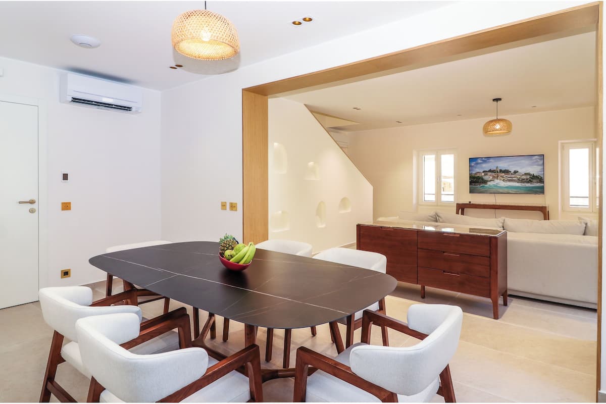 Un comedor moderno con una mesa de madera oscura y sillas blancas tapizadas, un mueble de madera y un cuadro sobre un sofá.