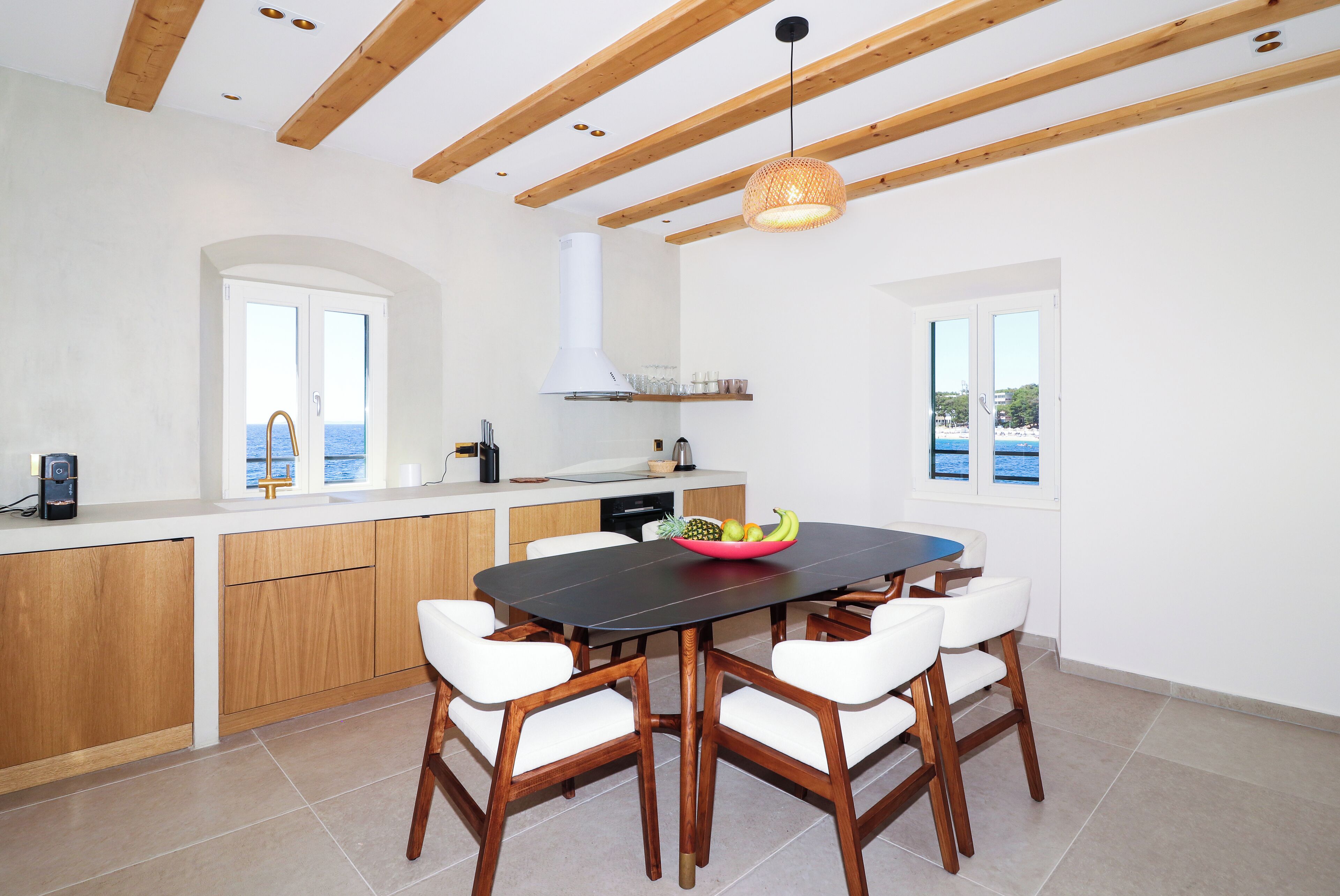 A modern kitchen with wooden beams, a dining table with white chairs, and a view of the sea through large windows.
