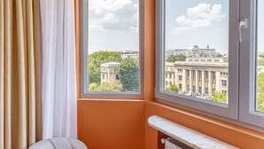 A corner room with an orange wall, a large window with a view of a building, a radiator, and a grey chair. - Playful Bright 1BR Near The Faculty of Law (Bucharest)
