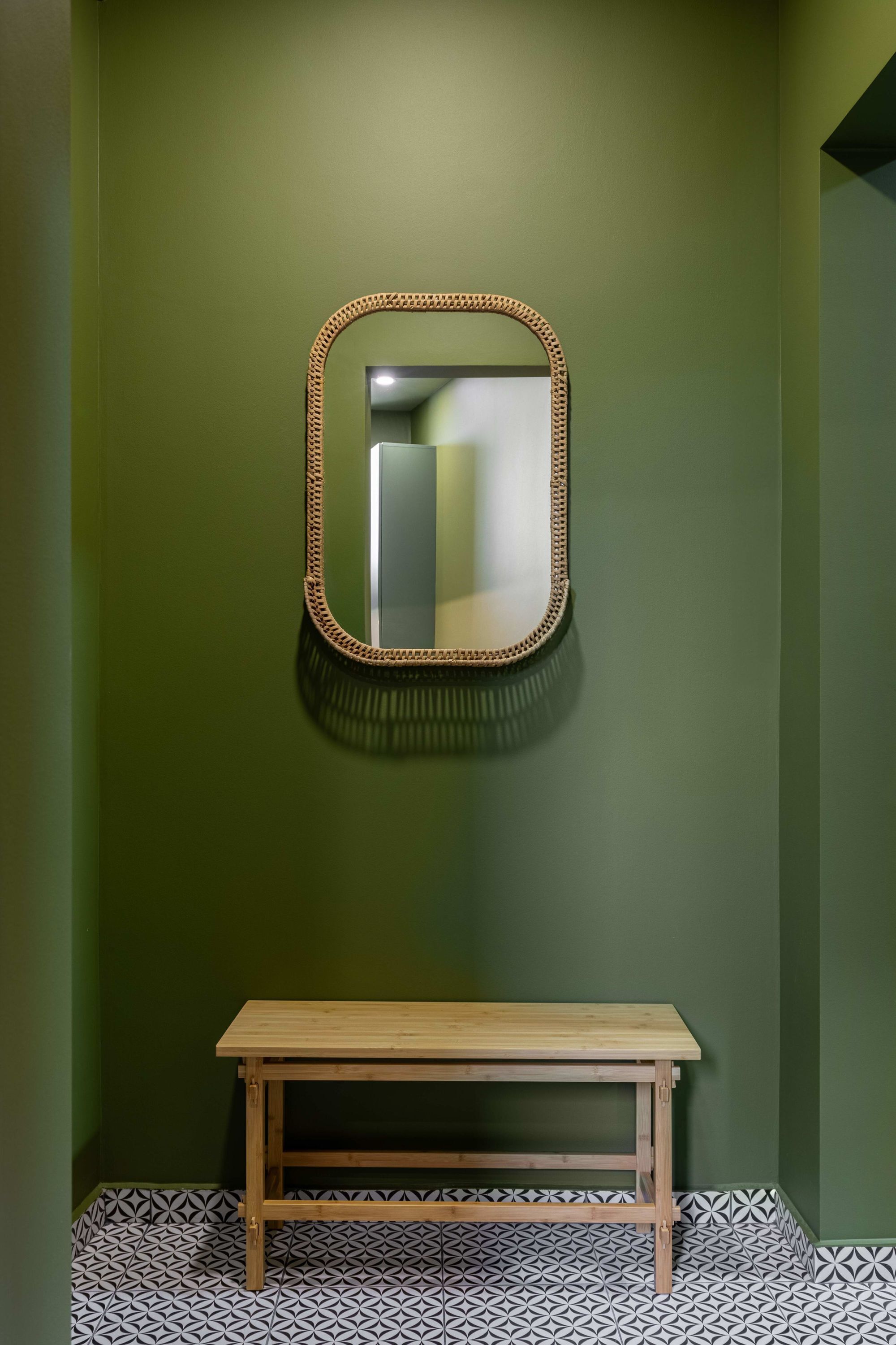 Un baño con pared verde, un banco de madera y un espejo con marco de mimbre.