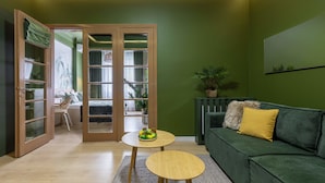 Una habitación con pared verde, un sofá verde con un cojín amarillo, una mesa de madera con un frutero y una silla de madera.