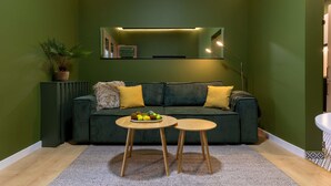 Una habitación con paredes verdes, un sofá verde oscuro, dos mesas redondas de madera, una planta en maceta y un espejo sobre el sofá.