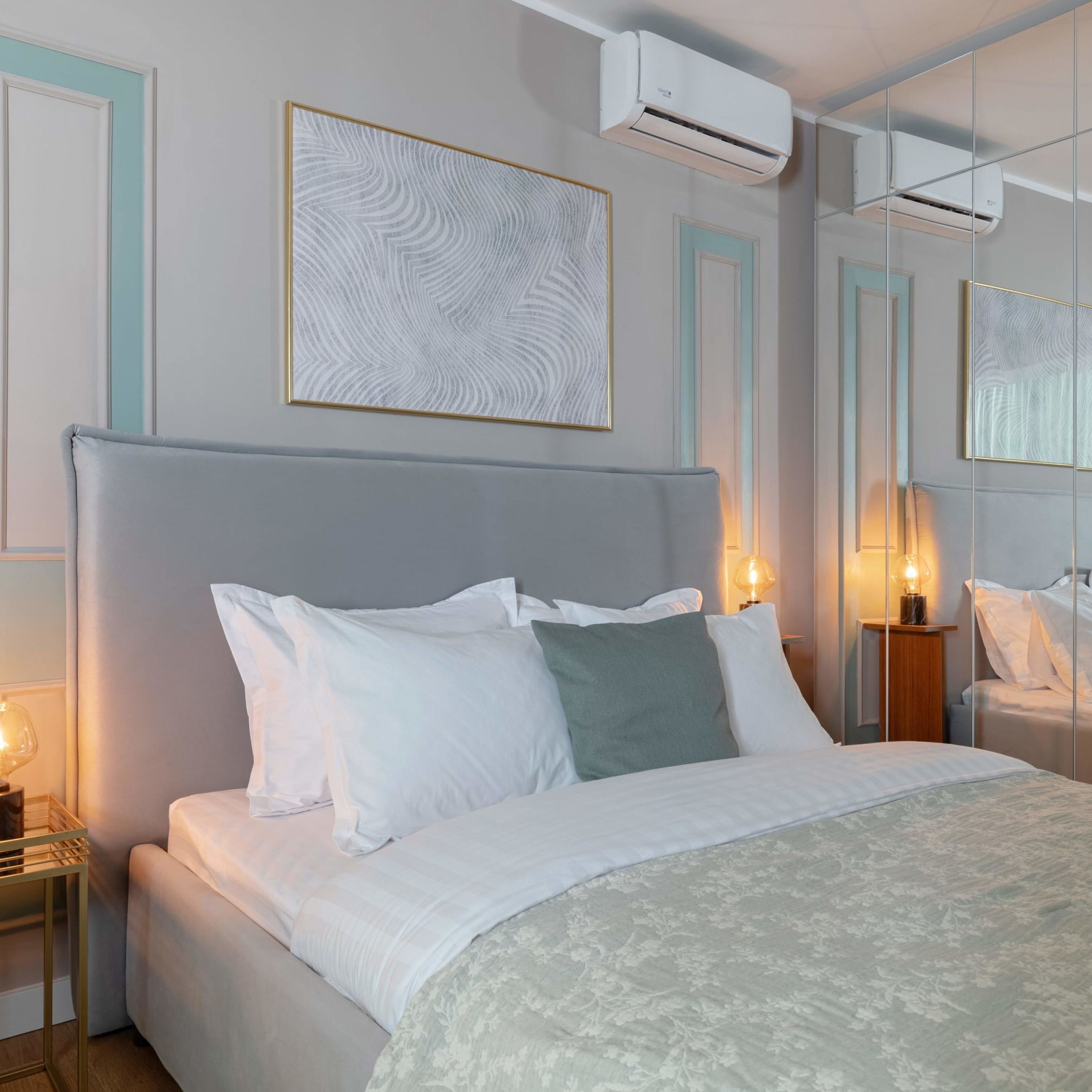 A neatly made bed with white and green pillows, a grey headboard, and a patterned bedspread. There are bedside lamps and a wall-mounted air conditioner.