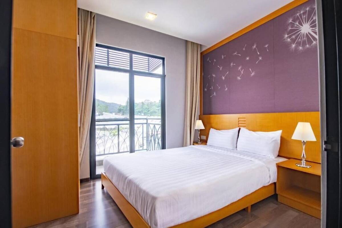 A hotel room with a large bed, wooden headboard, two bedside tables with lamps, a view of greenery through a large window, and a balcony.