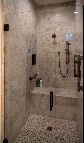Eine begehbare Dusche mit beigen Fliesen, einer Handbrause, einem am Wand montierten Wasserhahn und einem Ablagebereich für Pflegeprodukte.