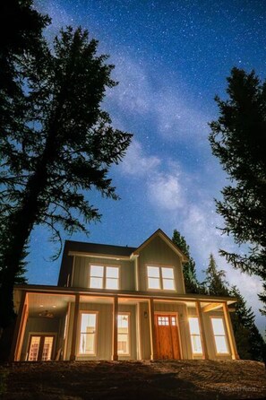Ein zweistöckiges Haus mit vielen Fenstern und einer Holztür, umgeben von Bäumen unter einem sternenübersäten Nachthimmel.
