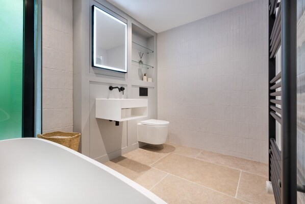Un baño moderno con bañera, inodoro y un estante de vidrio con artículos de aseo.