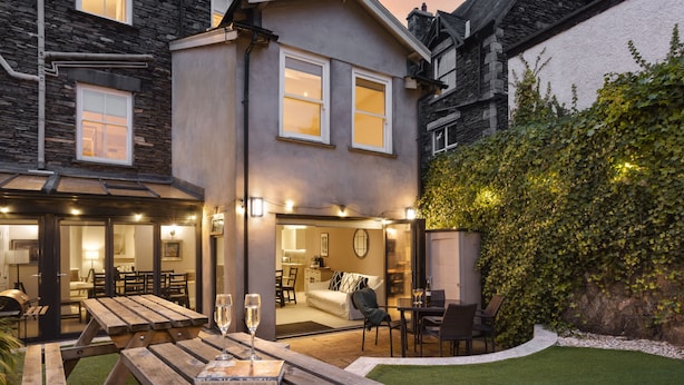 A modern house with a patio area featuring a wooden table and chairs, a small garden, and a view of the interior.