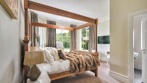 A four-poster bed with a fur throw, a lamp, and a view of the outdoors through large windows.