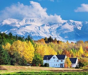 A white house with a dark roof, surrounded by autumn-colored trees and a snow-capped mountain in the background.