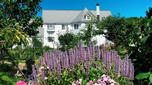 Een wit huis met een grijs dak, omringd door weelderige tuinen met roze en paarse bloemen.