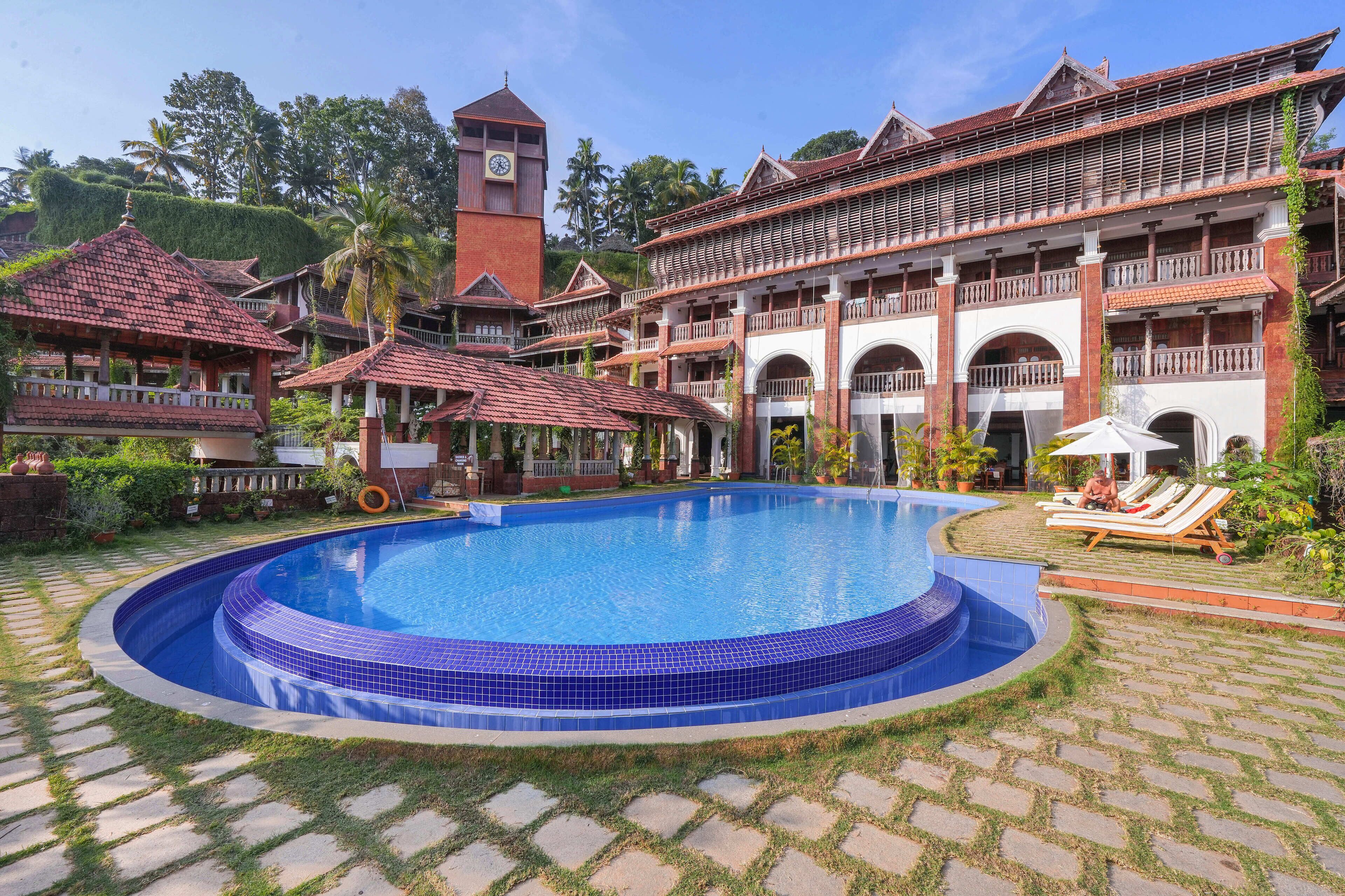 A swimming pool surrounded by a traditional building with a clock tower, palm trees, and a well-maintained lawn.
