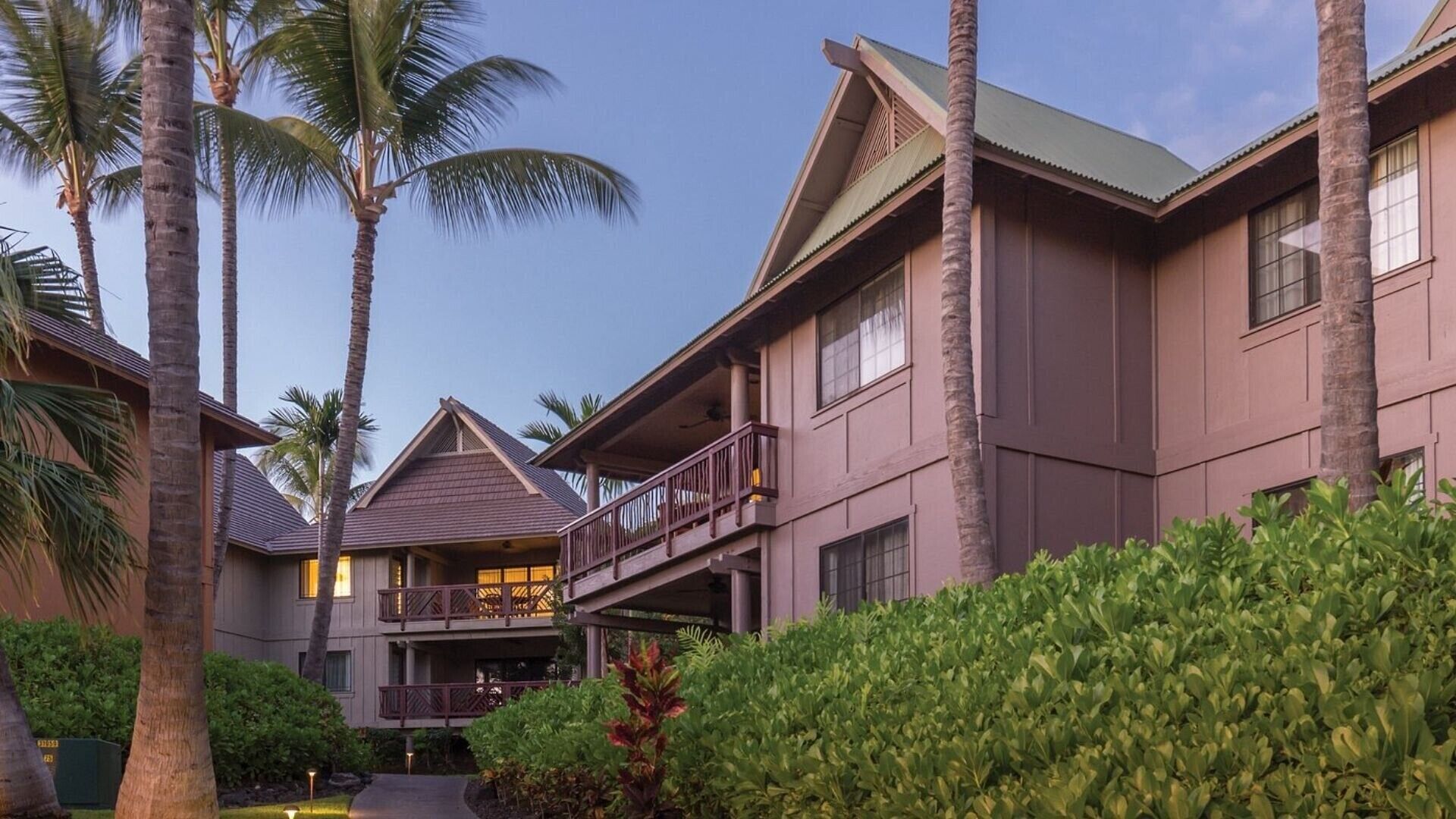 A multi-story hotel with a balcony on the second floor, surrounded by palm trees and well-maintained greenery.