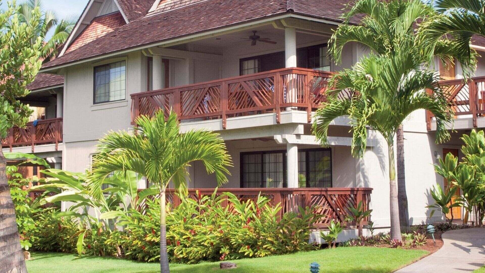 Un hôtel de deux étages doté d’un balcon au deuxième étage, entouré de plantes tropicales et d’une pelouse bien entretenue.