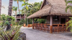 A resort with a thatched-roof bar area, wooden chairs, and a view of palm trees and a building with balconies.