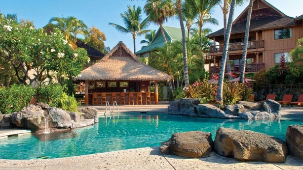 A pool area with a thatched-roof bar, palm trees, and a multi-story building with balconies.