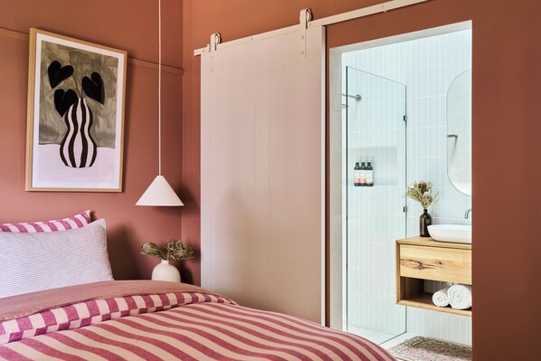 Un dormitorio con cama, una mesita de noche con un florero, una obra de arte enmarcada, y una puerta corredera que da a un baño con espejo, lavamanos y toallero.